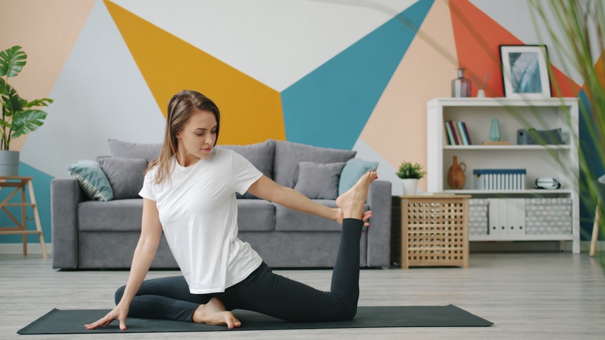 Cute blonde in leggings and t-shirt is stretching legs doing yoga at home on mat caring for body health in modern apartment. Lifestyle and sports concept.